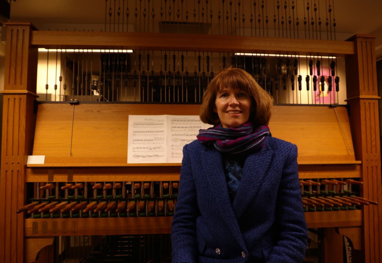A-woman-with-a-short-bob,-scarf,-and-blue-blazer-sits-in-front-of-a-carillon-instrument.
