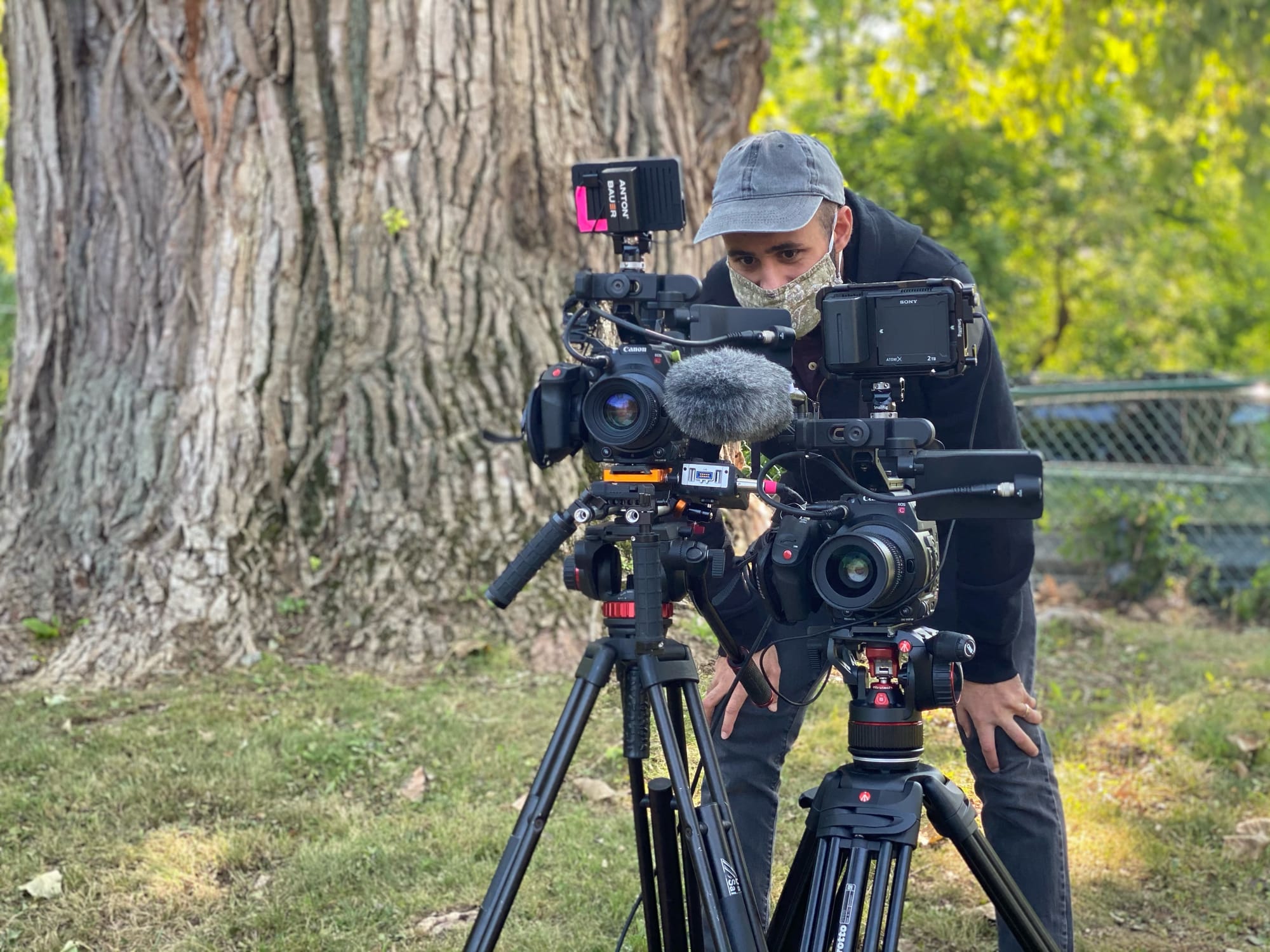 A man in a baseball cap and face mask stands behind two large cameras on tripods. 