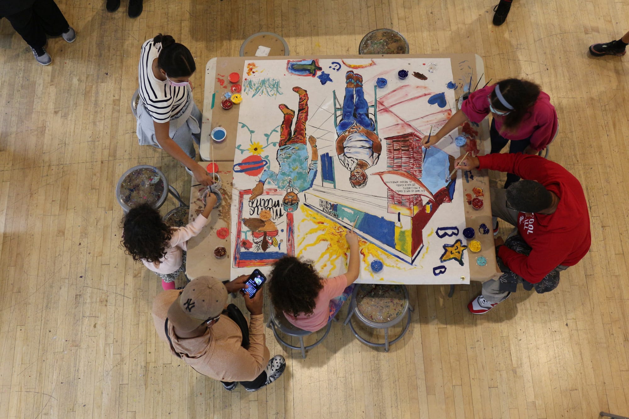 An overhead shot of a group of 6 people, a mix of kids and adults sitting at a table and working on a painting.