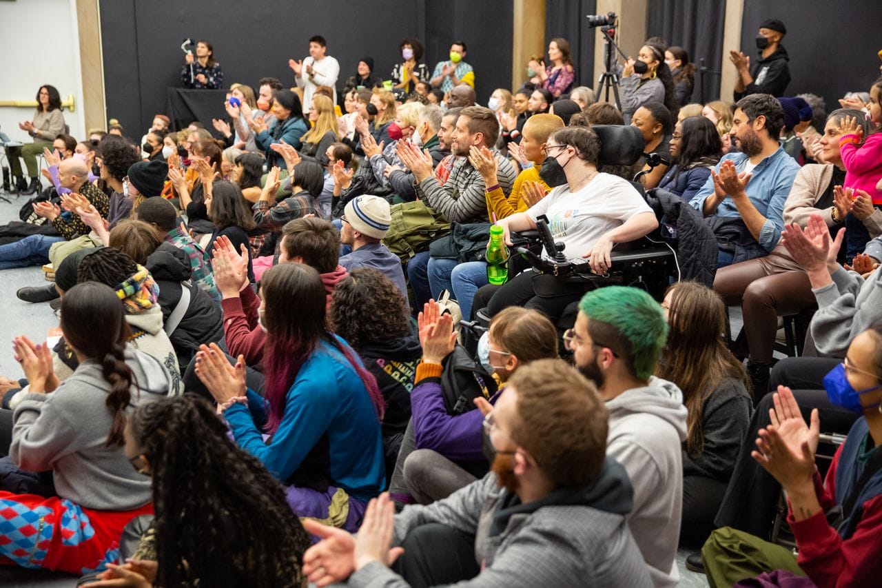 A crowd of people sit and watch a performance and clap