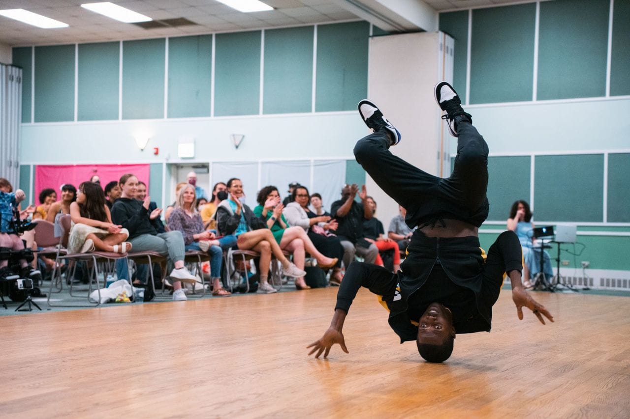 A dancer balances on his head on a wooden stage while an audience looks and claps.