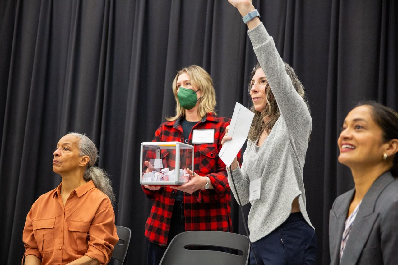 Four women are in this photo, two sitting and two standing. One woman dressed in a red plaid shirt holds a clear box filled with slips of white paper