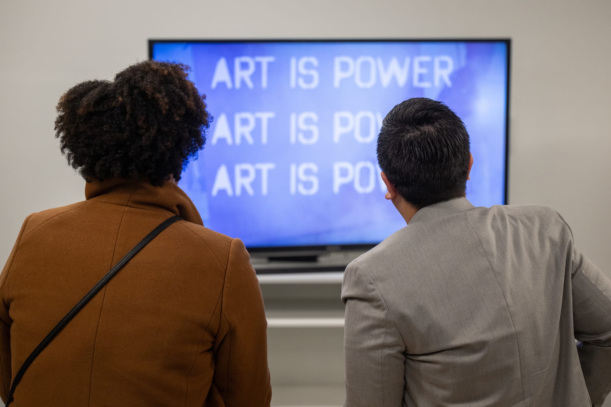 Two people, photographed from behind watch a video on a television. The tv shows the quote, Art is Power