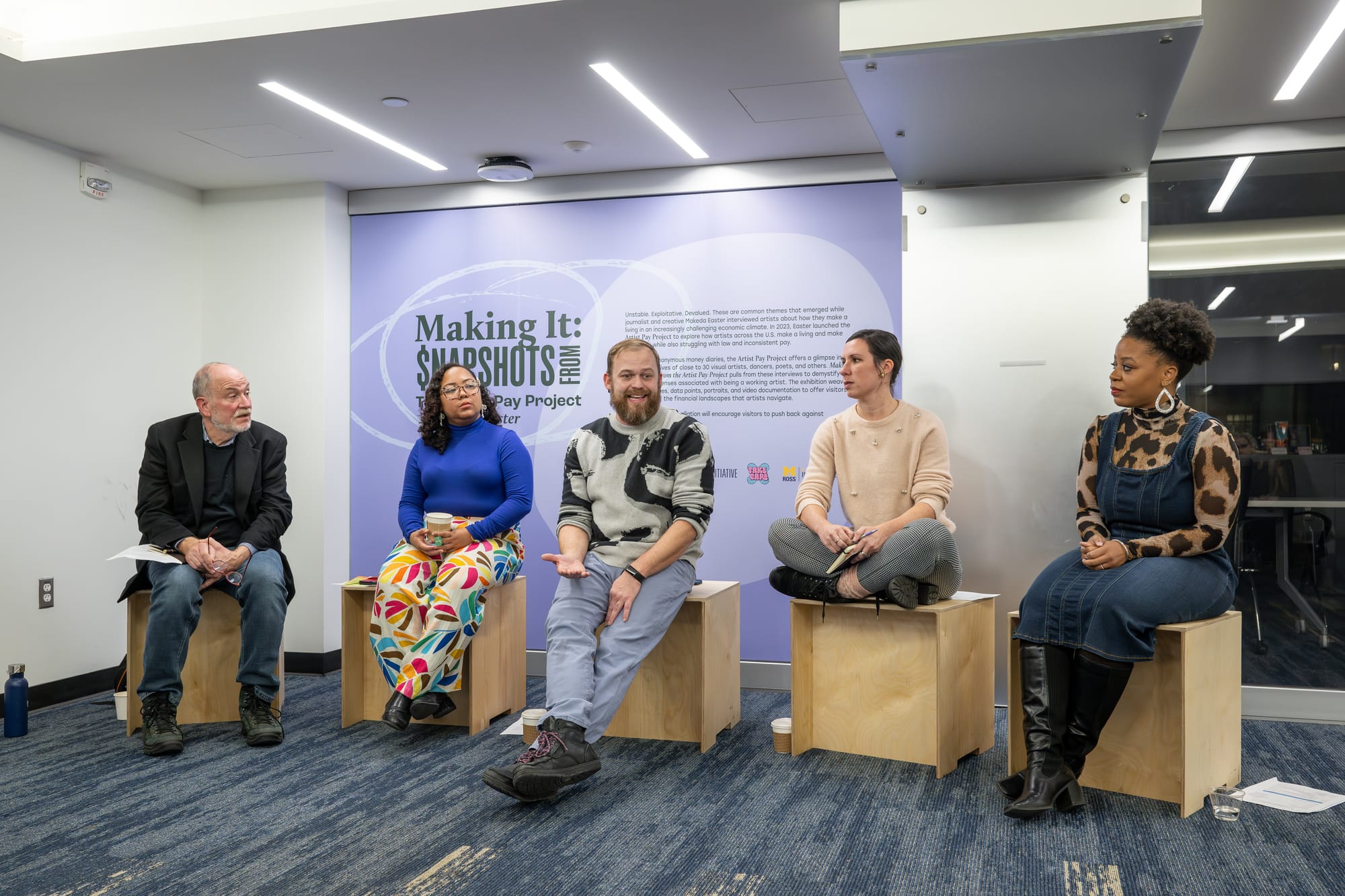 Five people sit on wooden benches in the installation space. They're speaking on a panel