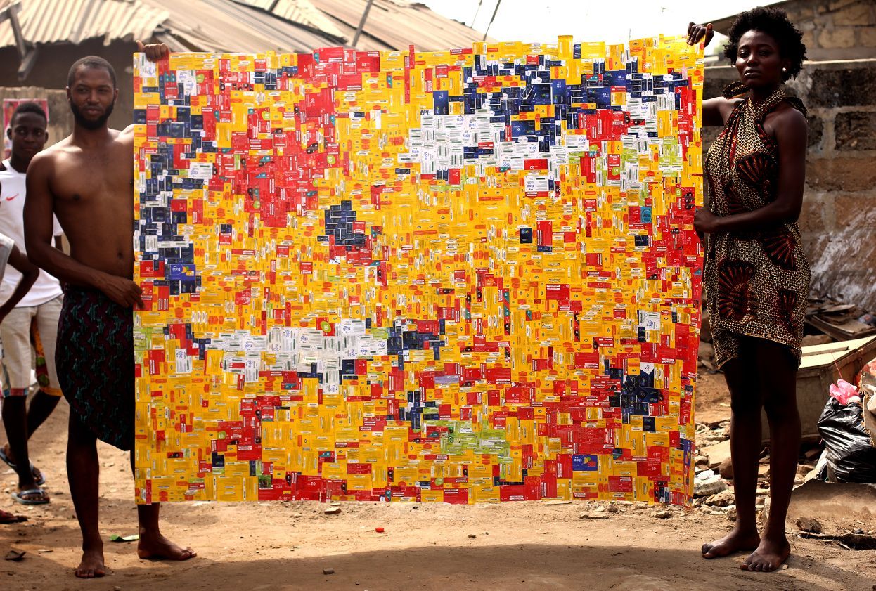Basil-Kincaid-holds-up-a-collaged-art-work-made-of-yellow,-red,-and-white,-pre-paid-calling-cards-with-a-collaborator-in-Ghana.