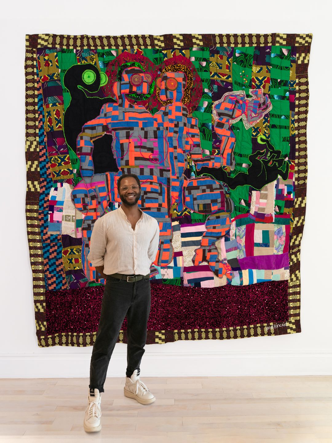 Artist-Basil-Kincaid,-dressed-in-jeans-and-a-button-down-shirt-stands-in-front-of-one-of-their-colorful-quilts.-