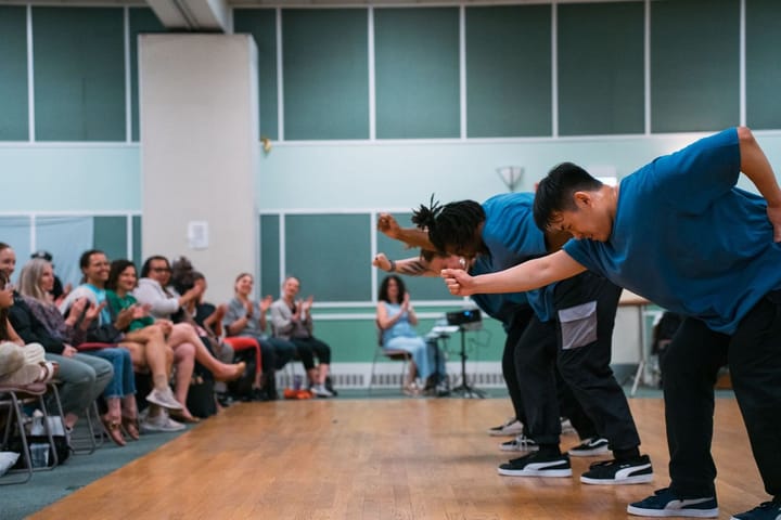 3 dancers wearing blue shirts perform for an audience. Their arms are outstretched and they're in line.